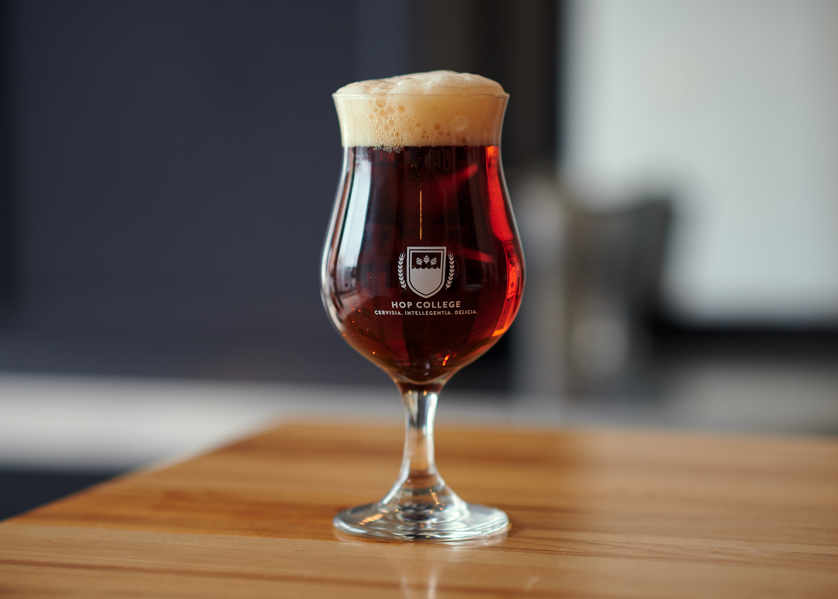 GLBC Hop College tulip glass filled with an amber colored beer on a wooden bar top.