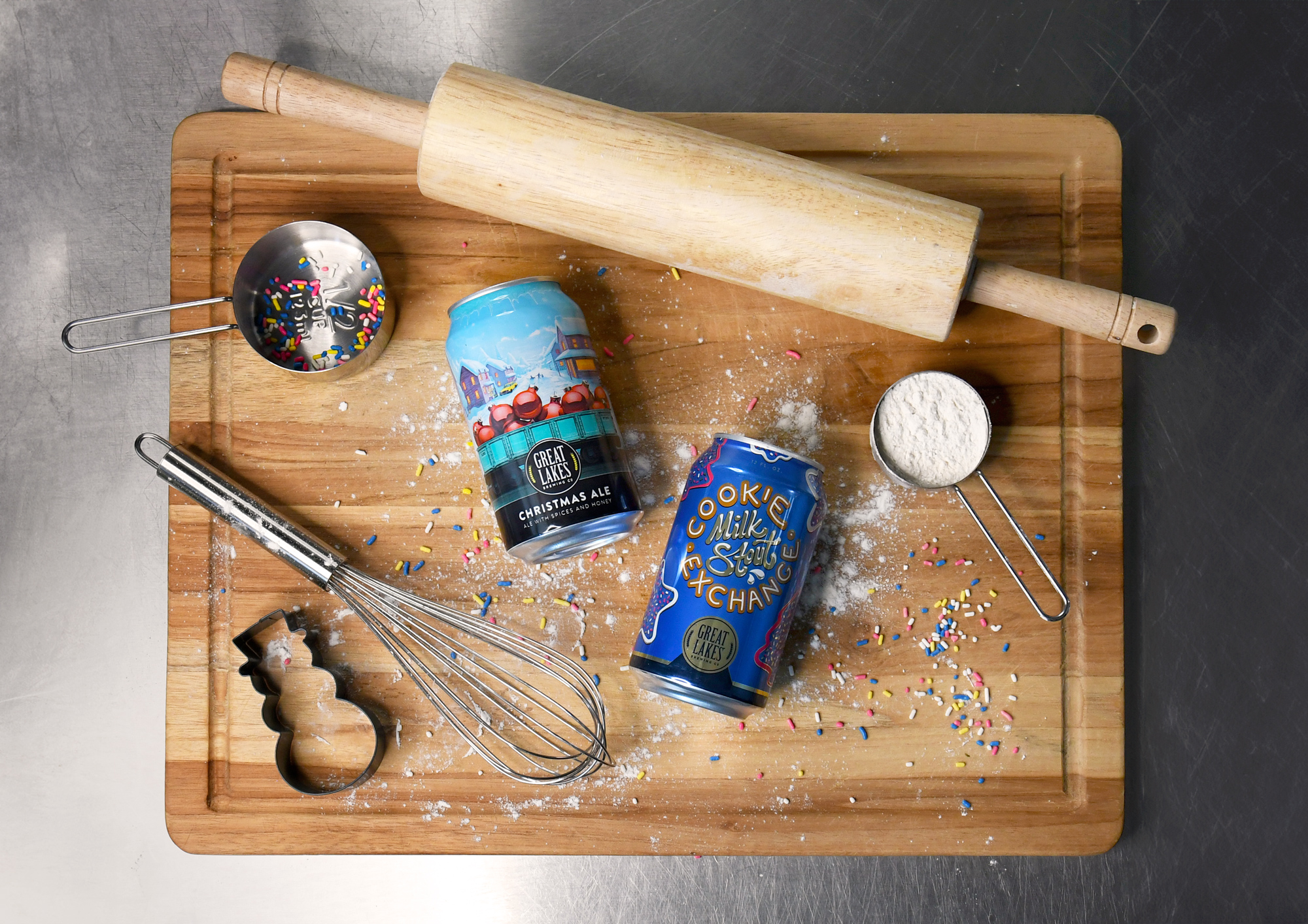 Cans of Cookie Exchange Milk Stout and Christmas Ale laid on a cutting board with baking utensils.