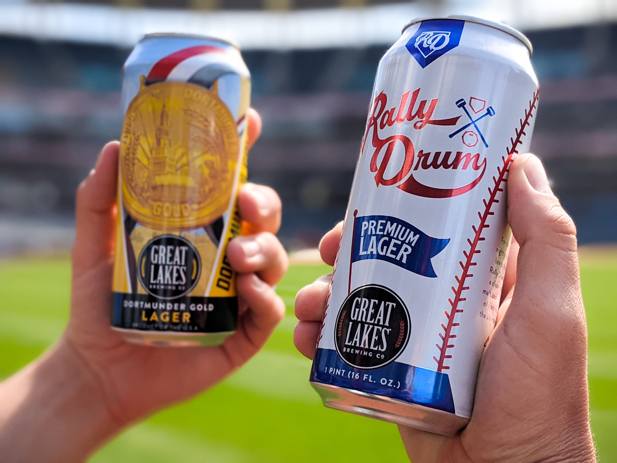 Hands holding 16 oz. Cans of Rally Drum Premium Lager and Dortmunder Gold Lager in front of a baseball field.