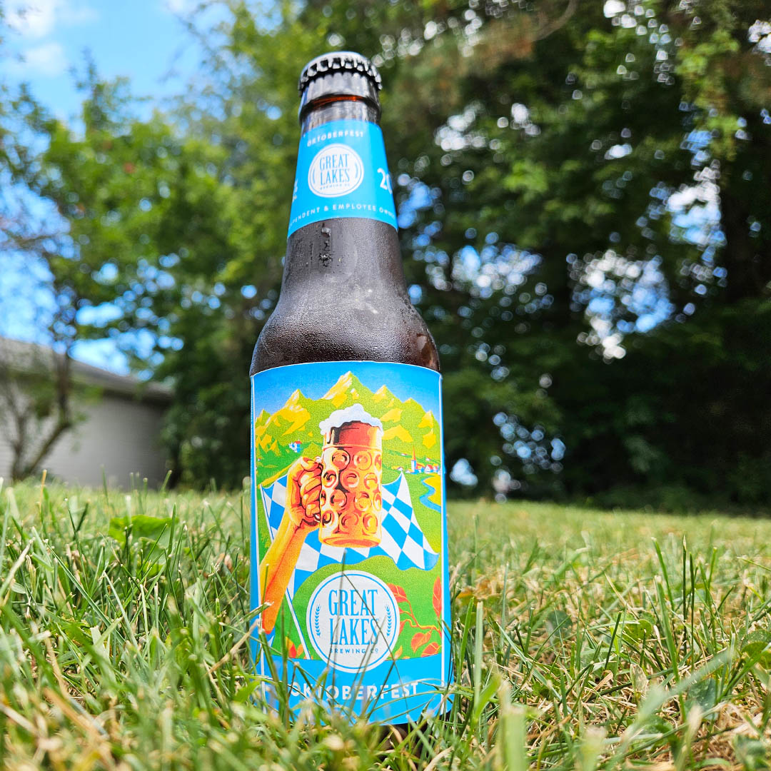 12 oz. Bottle Oktoberfest sitting in green grass with a white shed, leafy trees, and a bright blue sky in the background.