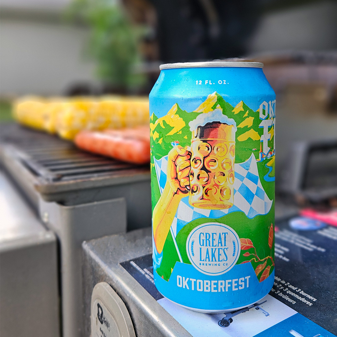 12 oz. Can Oktoberfest placed on the side of a grill, with hot dogs and corn on the cob on the griddle