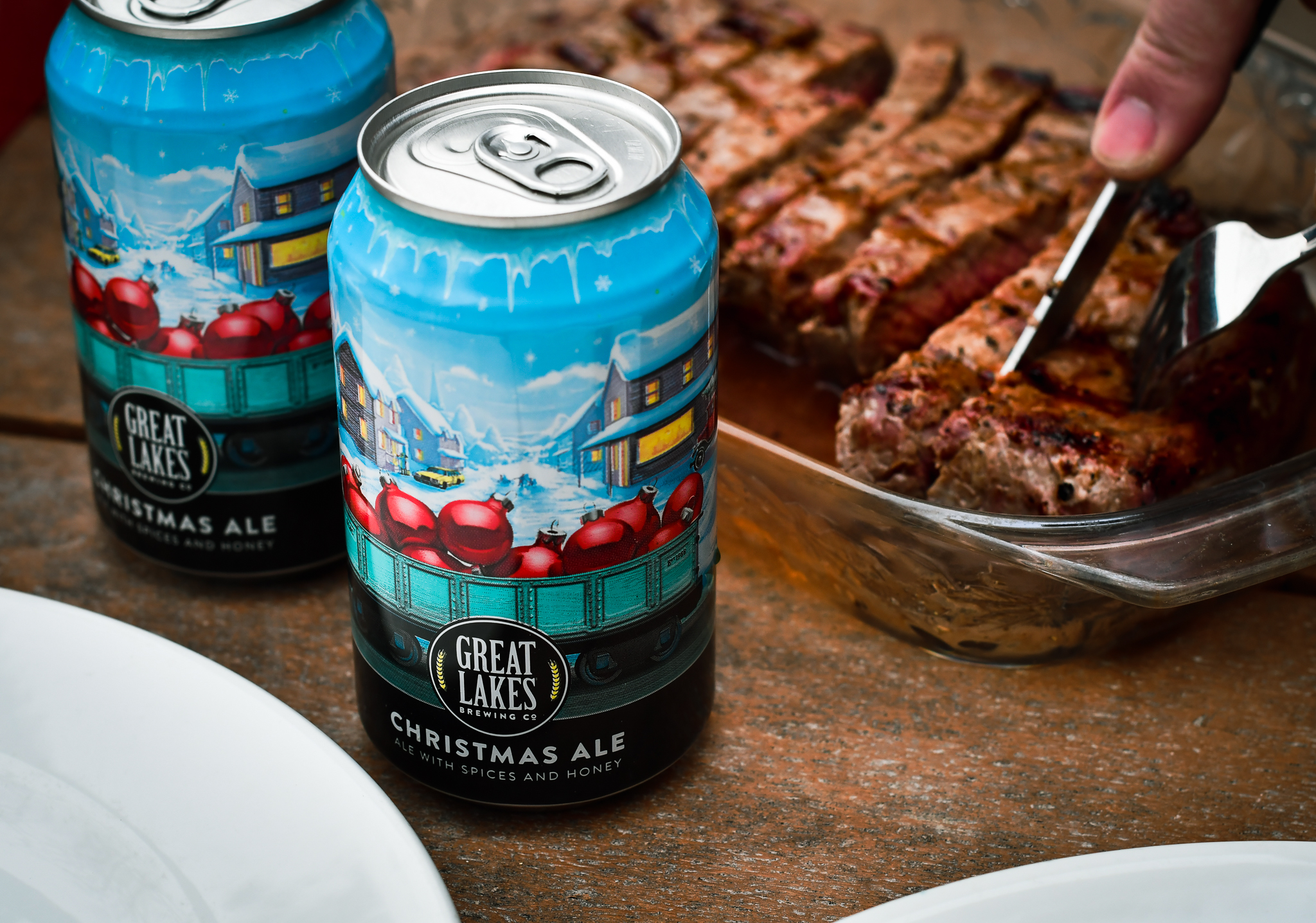 Two cans of Christmas Ale in front of a person cutting a steak.