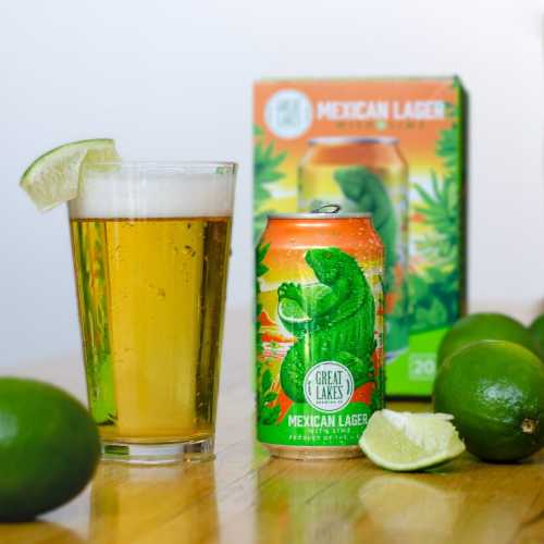 Pint glass filled with Mexican Lager with Lime, garnished with a lime wedge, sitting next to a can, 6-pack, and full limes.