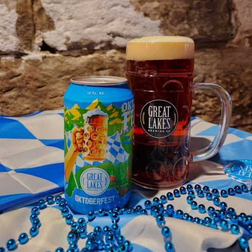 12 oz. Can Oktoberfest next to a stein of the beer, laid upon a Bavarian flag and next to a set of blue beads.